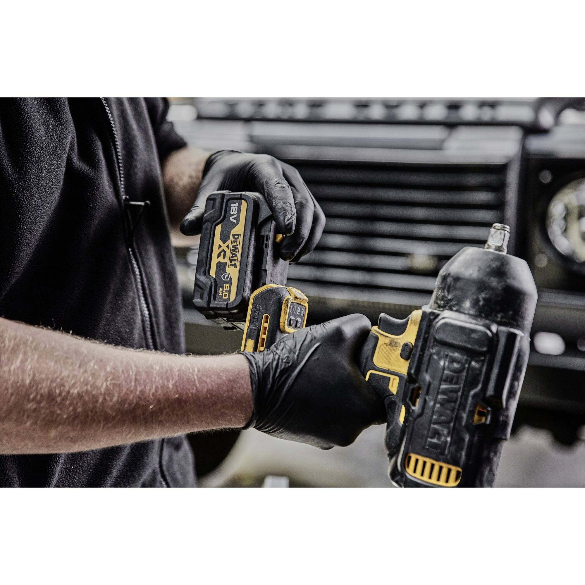 A person is holding an electric impact driver with yellow and black details, with a vehicle standing in the background.