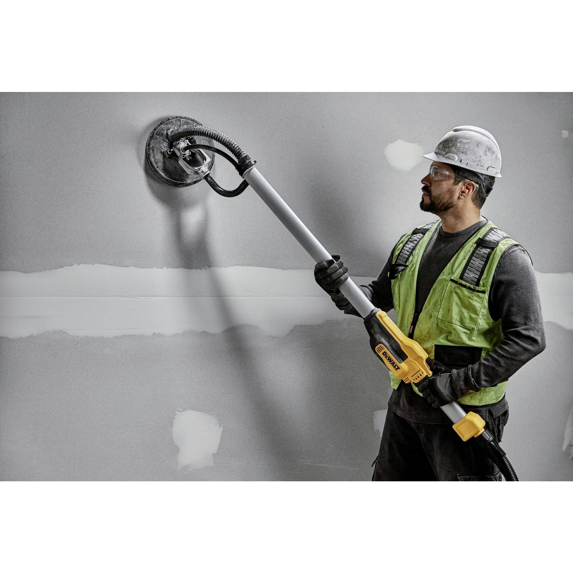 A construction worker wearing a hard hat and high-visibility vest is grinding a wall inside a room using a long grinding machine.