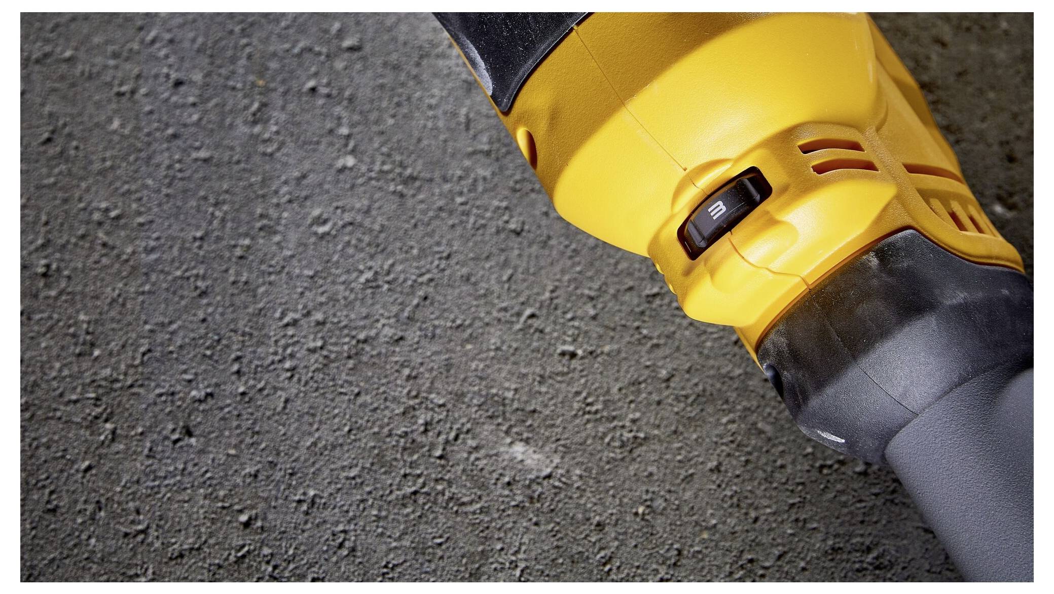 A yellow electrical power tool lies on a grey, rough concrete floor. The focus is on a black switch on the device.