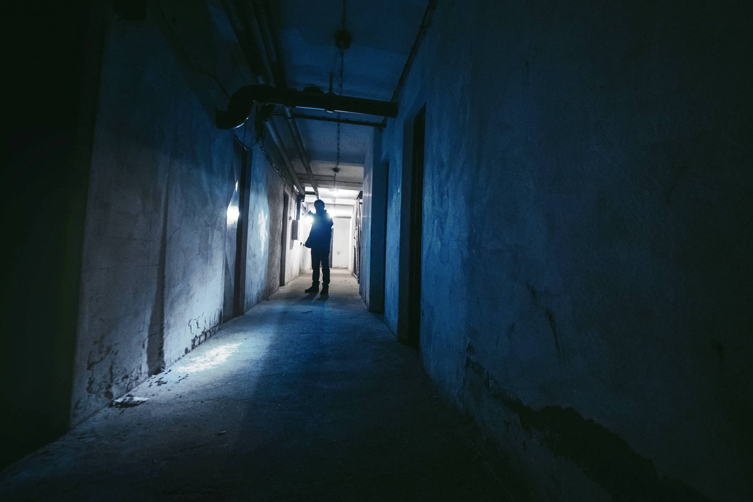 A person stands at the end of a long, dark corridor, illuminated by a blue light source. The atmosphere feels mysterious.
