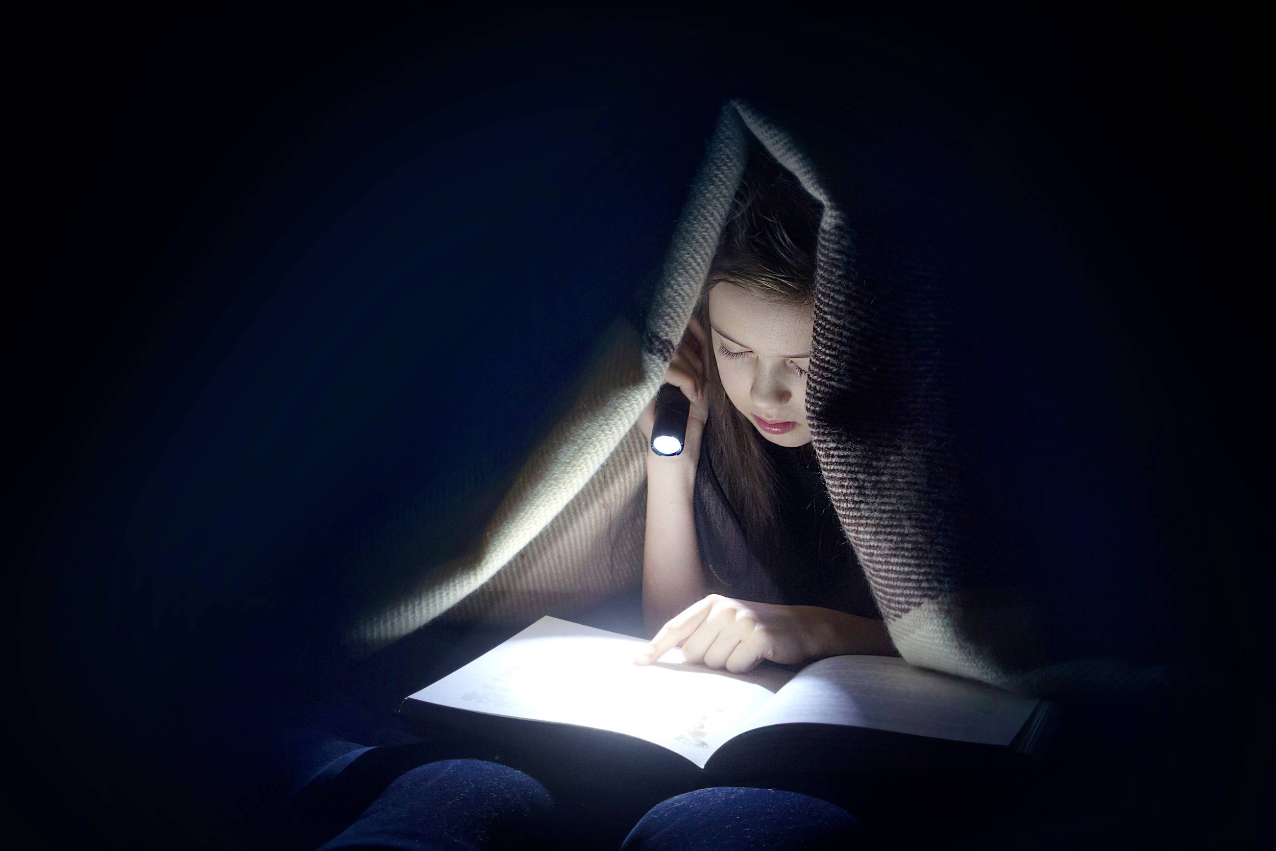 A child is reading a book under a blanket at night with a torch. The surroundings are dark, with the face visible in the torchlight.