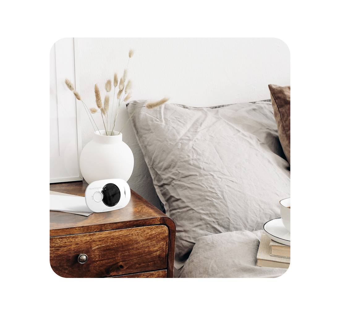 A bedside table with a modern surveillance camera, a vase with dried flowers, books and a mug in a cosy bedroom.