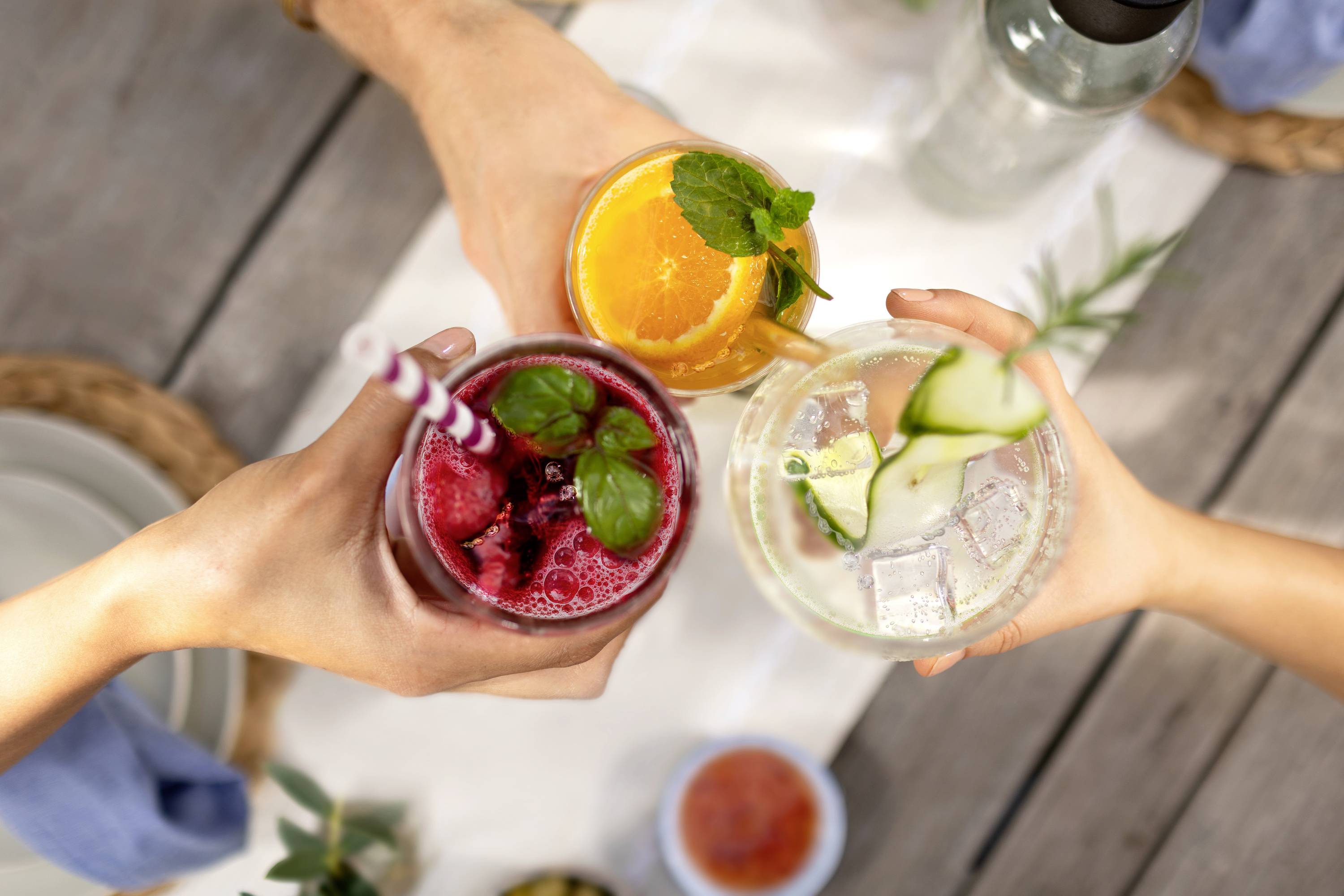 Three people toast with colourful cocktails, decorated with mint leaves, orange slices, and cucumber slices.