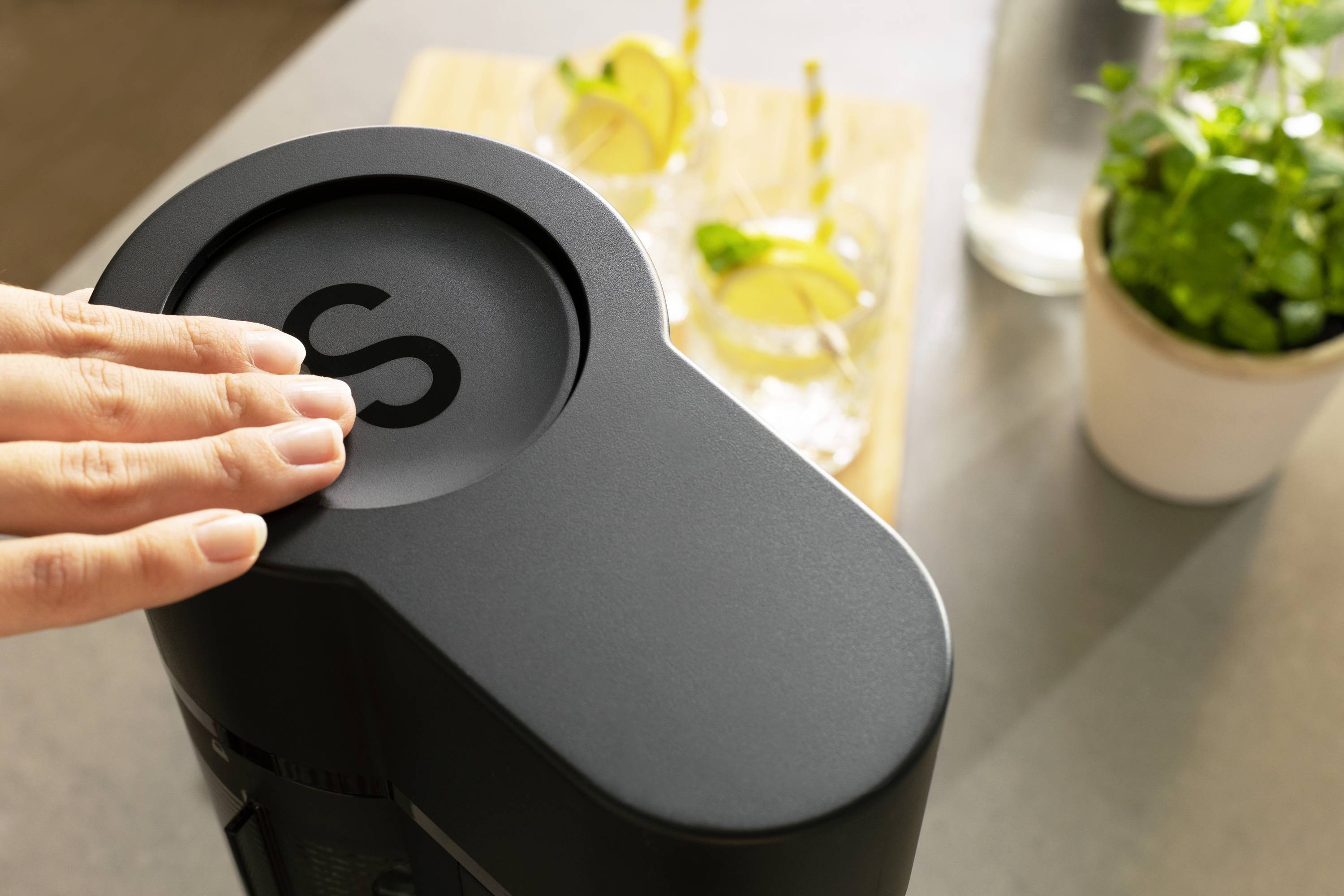 A hand operates a black sparkling water machine. In the background, drinks with lemon slices and a pot of herbs are visible.