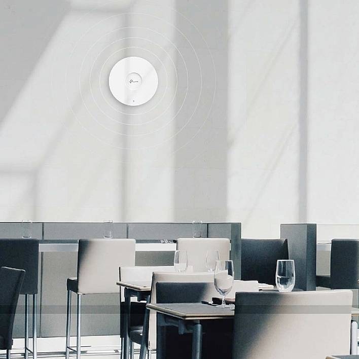 In a modern, bright conference room, a circular device with a logo hangs on the wall. Tables and chairs are ready.