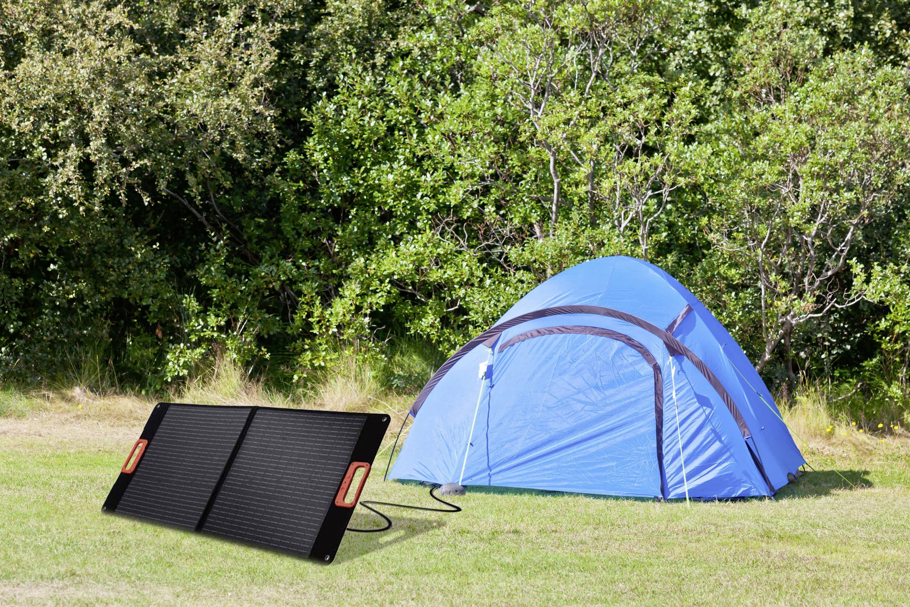 A blue tent is pitched on a meadow in front of a forest. Next to it lies a solar panel, used for generating electricity outdoors.