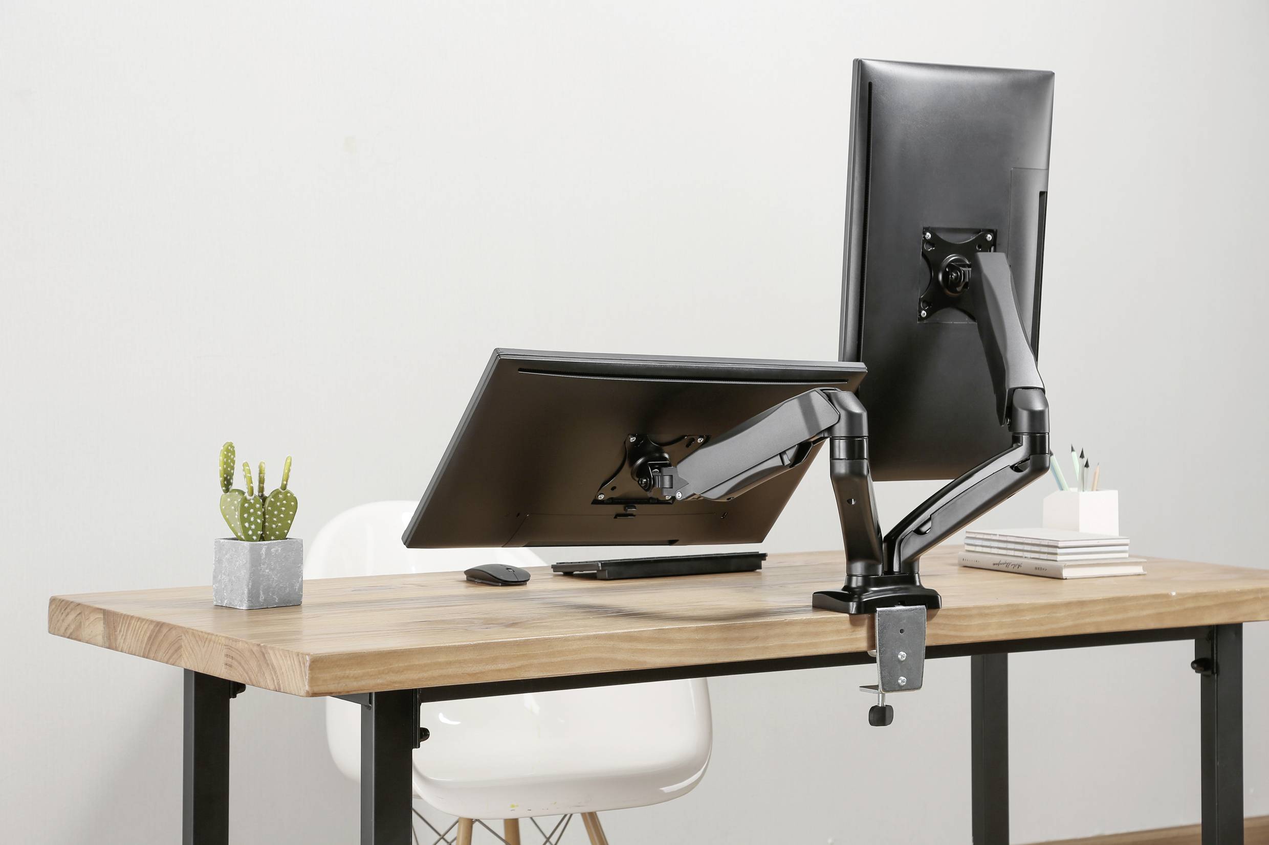 Two monitors on adjustable mounts on a wooden desk; one vertical, the other horizontal. A small plant on the left.