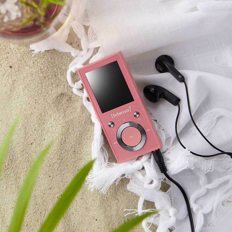 A pink music player with headphones is lying on a white cloth next to a drink and plants in the background.