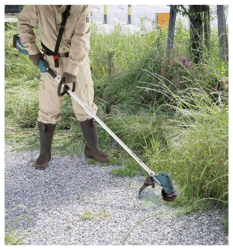 A person wearing tan protective clothing and boots is using a green and black string trimmer to cut grass along a gravel path.