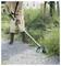 A person wearing tan protective clothing and boots is using a green and black string trimmer to cut grass along a gravel path.