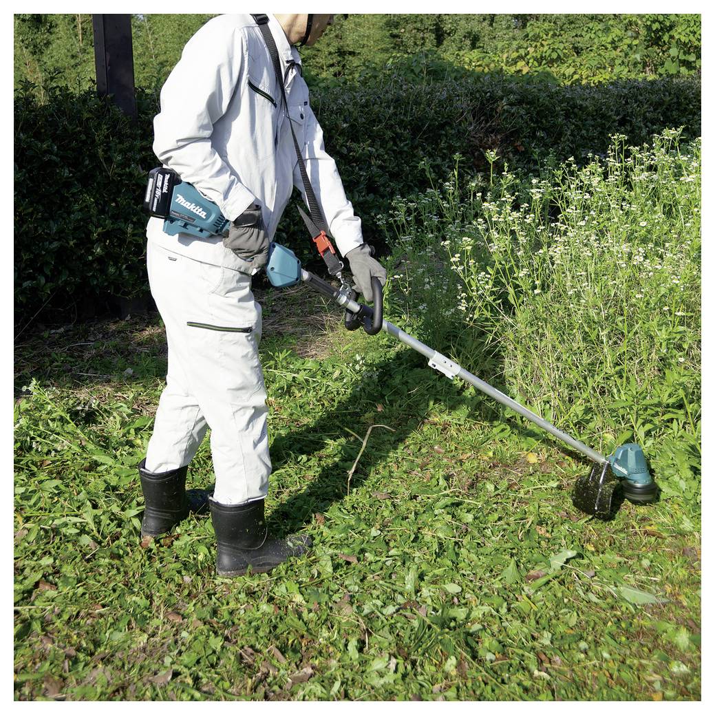 A person in protective clothing uses a grass trimmer to cut overgrown grass in a garden, surrounded by greenery.