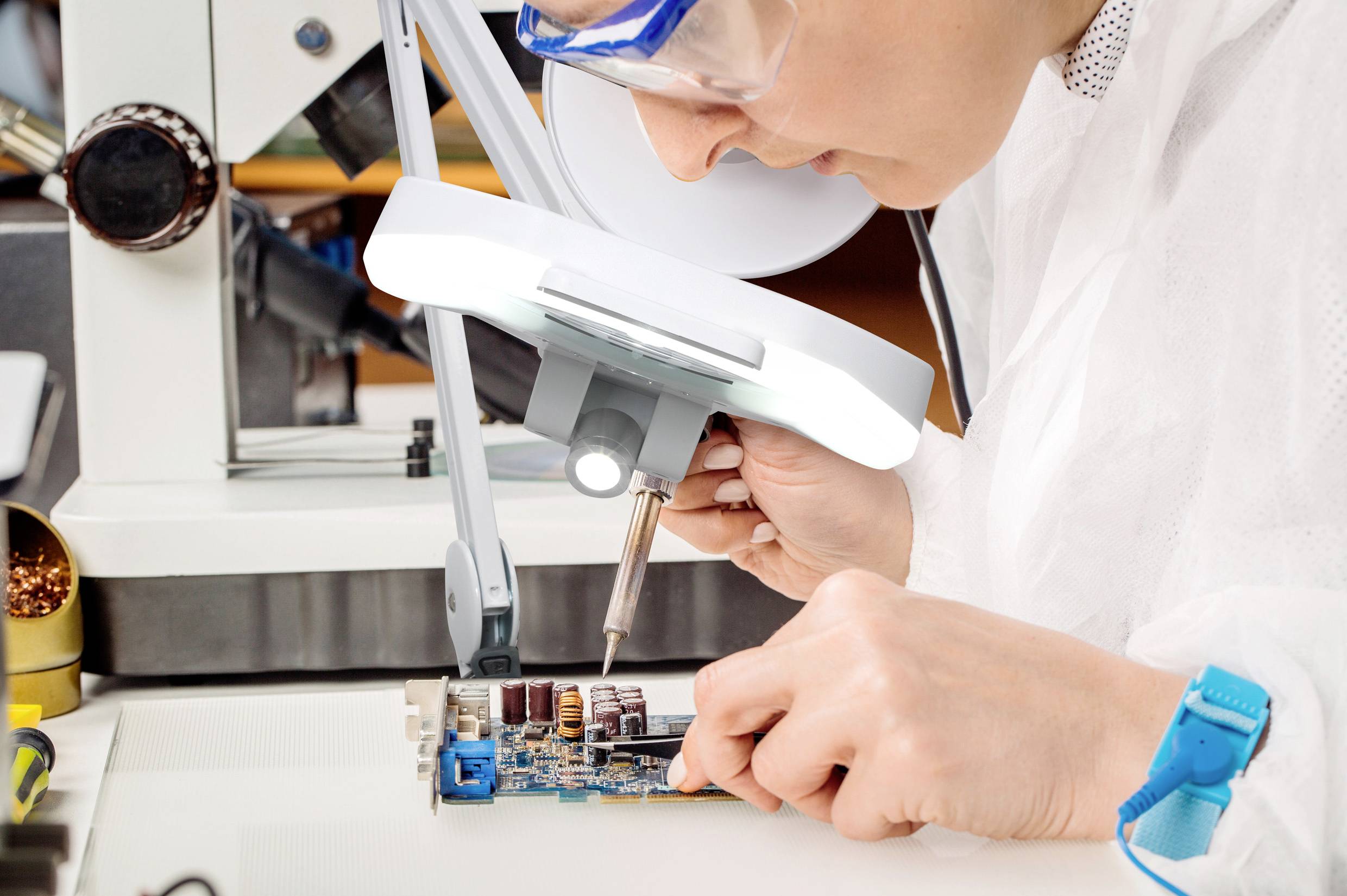 A person in a laboratory is working under a magnifying glass on a circuit board, using a soldering iron to secure components.