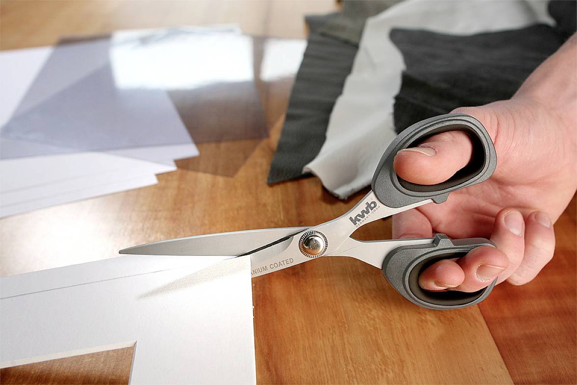 A hand is cutting white paper with scissors on a wooden table. Further pieces of paper and fabric are lying in the background.