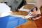 A hand is using large scissors to cut a blue sheet of paper on a wooden table. Fabric and paper are scattered in the background.
