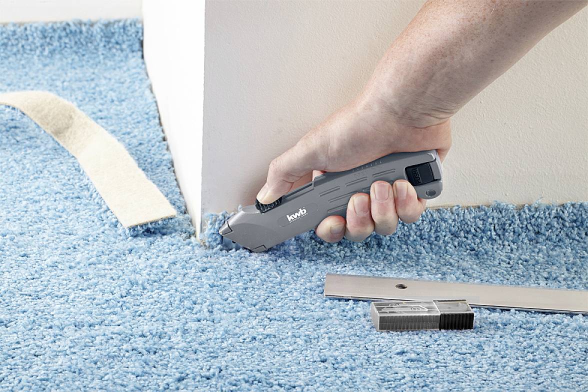 A hand is using a carpet knife to cut blue carpet along a white wall. A ruler and blades are lying nearby.