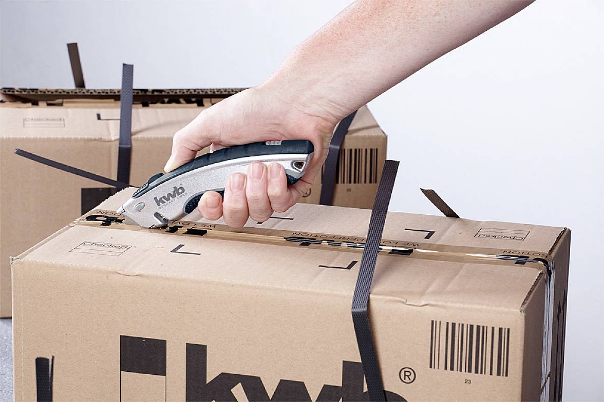 A hand uses a craft knife to cut through black plastic straps on a cardboard box. The box is ready for shipping.