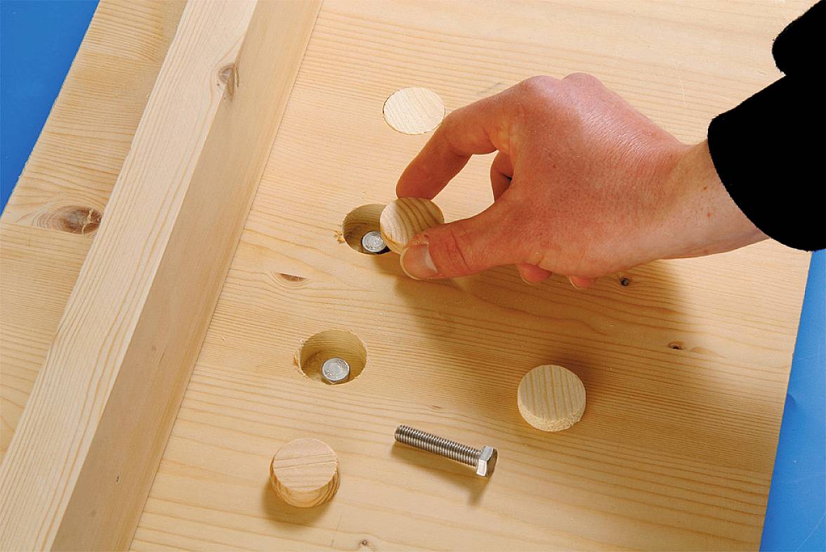 A hand places a wooden lid into a hole in a wooden board, with visible screws underneath.