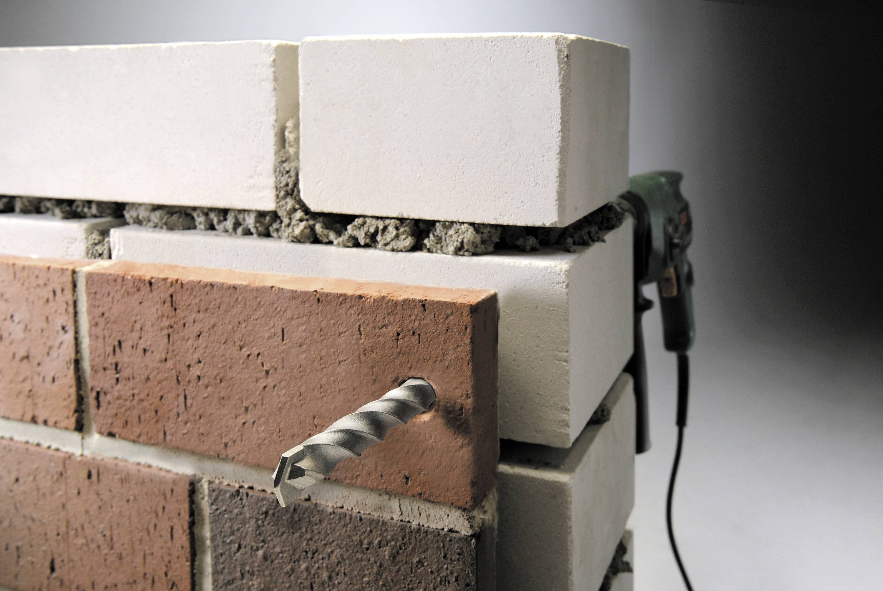 A drill is boring through a cross-layered wall composed of red and white bricks and grey mortar.