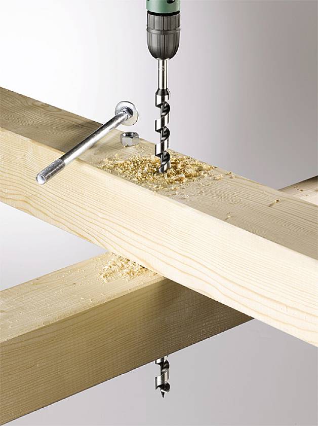 A drill pierces through two overlapping wooden beams. Wood shavings are scattered around the drill hole, with a spanner lying nearby.