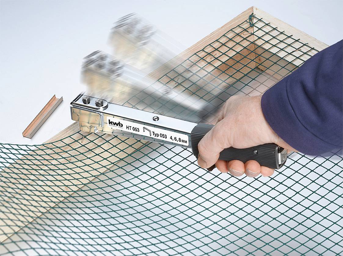 A hand is using a staple gun to secure a green mesh to a wooden frame. The stapler is moving quickly.