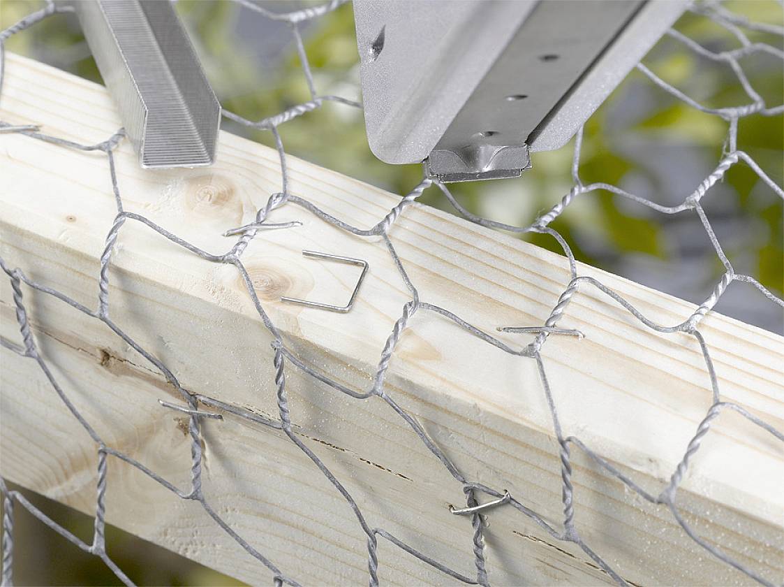 Chicken wire is secured to a wooden frame with clips. Blurred plants are visible in the background.