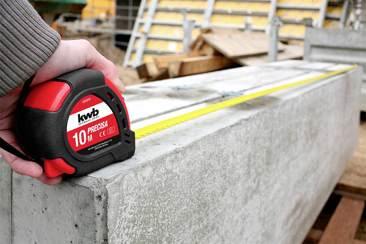 'Hand holding a tape measure over a concrete block with a yellow band, length measurement being carried out, construction materials in the background.'