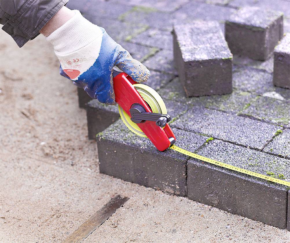 A person is measuring paving stones on a pavement using a tape measure. The paving stones are dark grey and the ground is sandy.