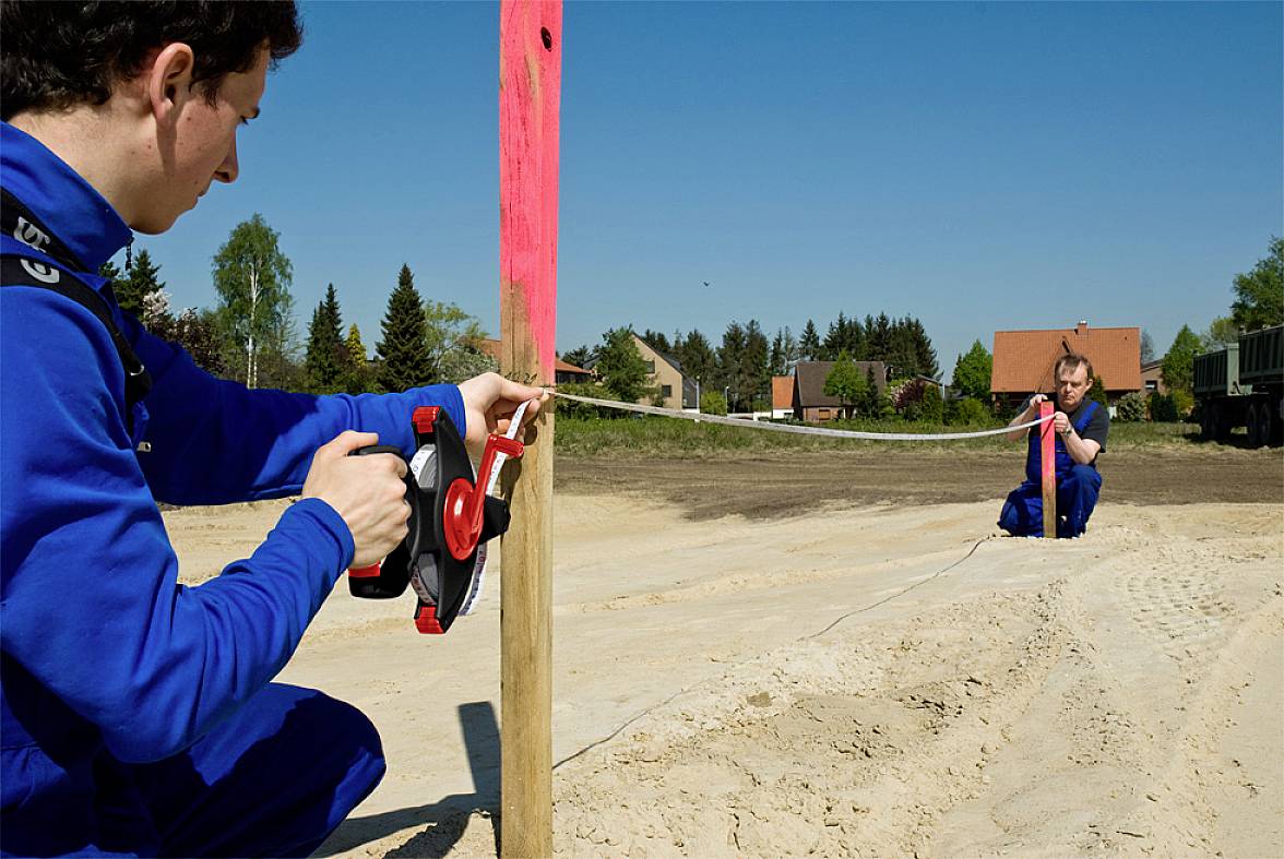 Two people in blue workwear are measuring the distance between a wooden post and a point on the ground at a construction site using a tape measure.