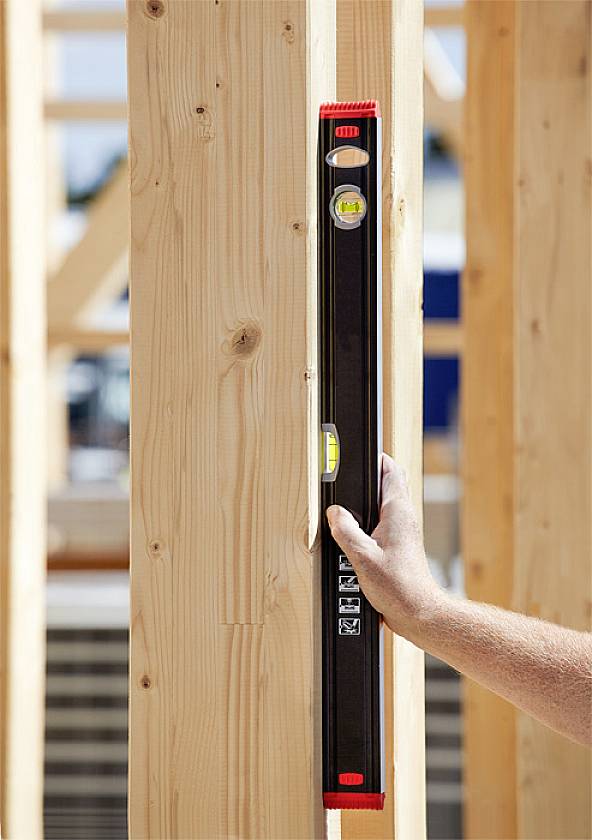 A person is holding a spirit level against a wooden post to check its verticality. The background shows timber frame construction.