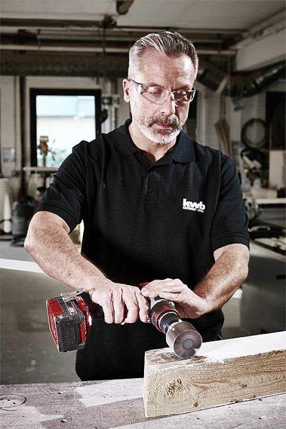 A man wearing safety glasses is using an electric drill to bore a hole into a piece of wood. A workshop is visible in the background.