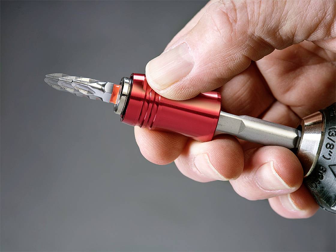 A hand is holding a red-metallic tool with a drill bit attachment, ready for use. In the background is a grey blurred area.