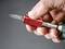 A hand is holding a red-metallic tool with a drill bit attachment, ready for use. In the background is a grey blurred area.