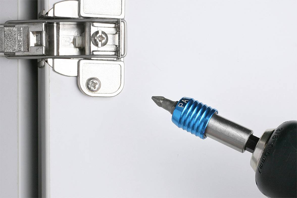 A blue magnetic bit holder secures a screw while a cordless drill inserts it into a metal hinge on a door.