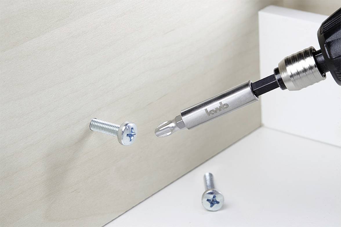 Cordless drill with screwdriver bit driving a screw into a wooden board. Another screw lies beside it.