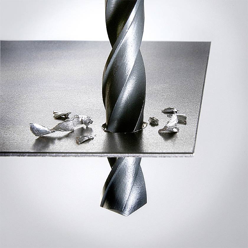 A metal drill bit pierces a thin metal sheet. Metal shavings are scattered around the drill hole.