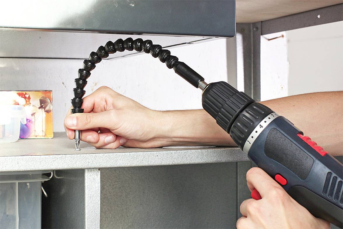 A person is using a cordless drill with a flexible drill extension to screw a fastener into a piece of furniture.