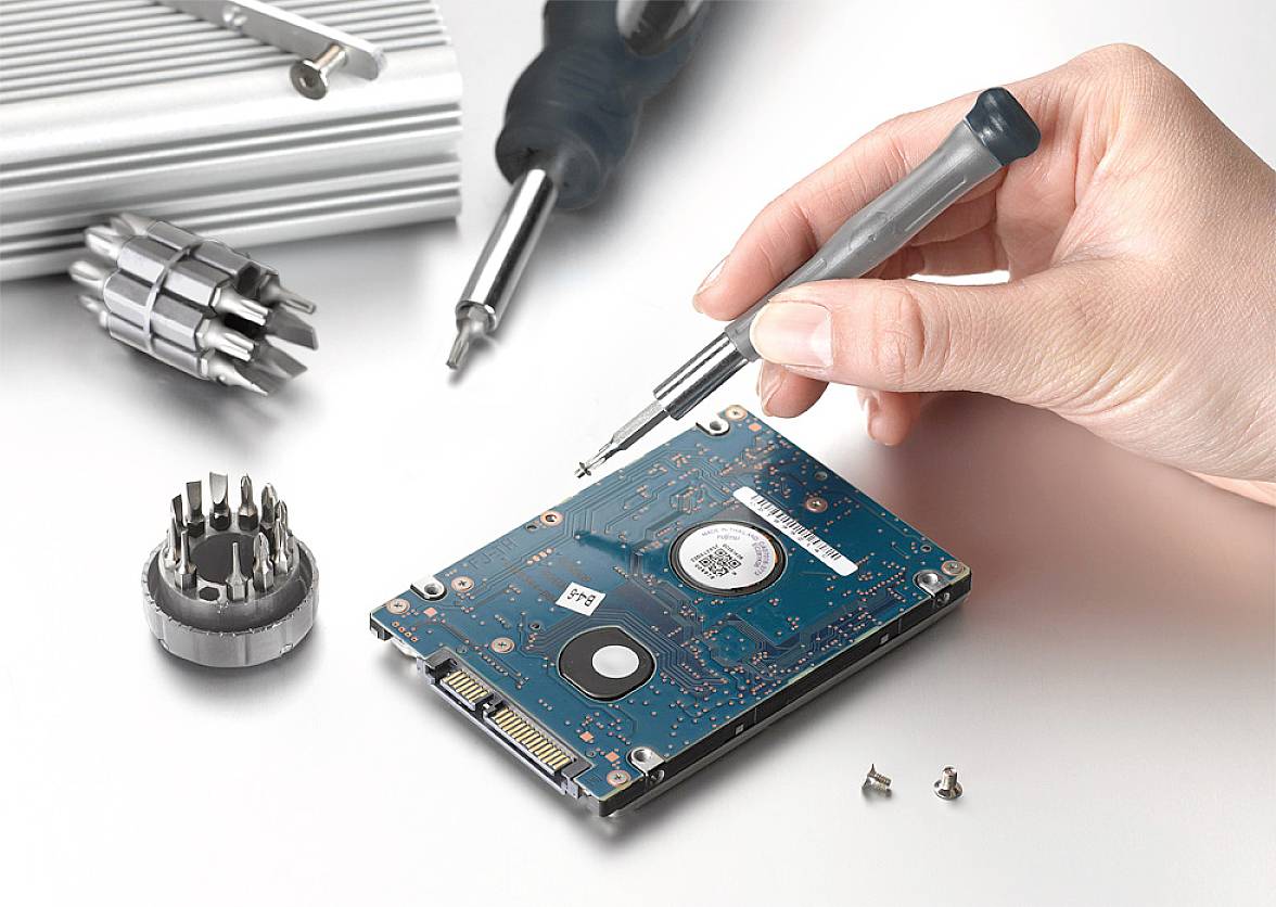 A hand holds a screwdriver over a hard drive. Various tools are scattered in the background.