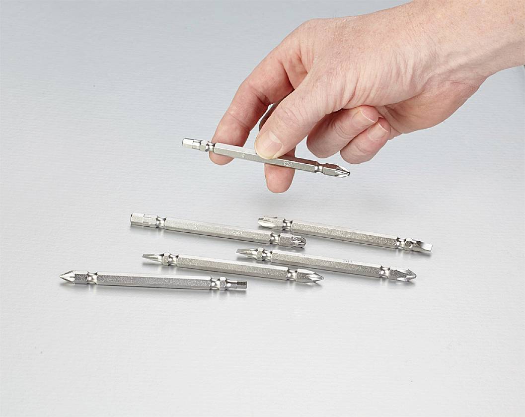 A hand is holding a double-sided screwdriver bit, similar to the five others lying on a light surface.