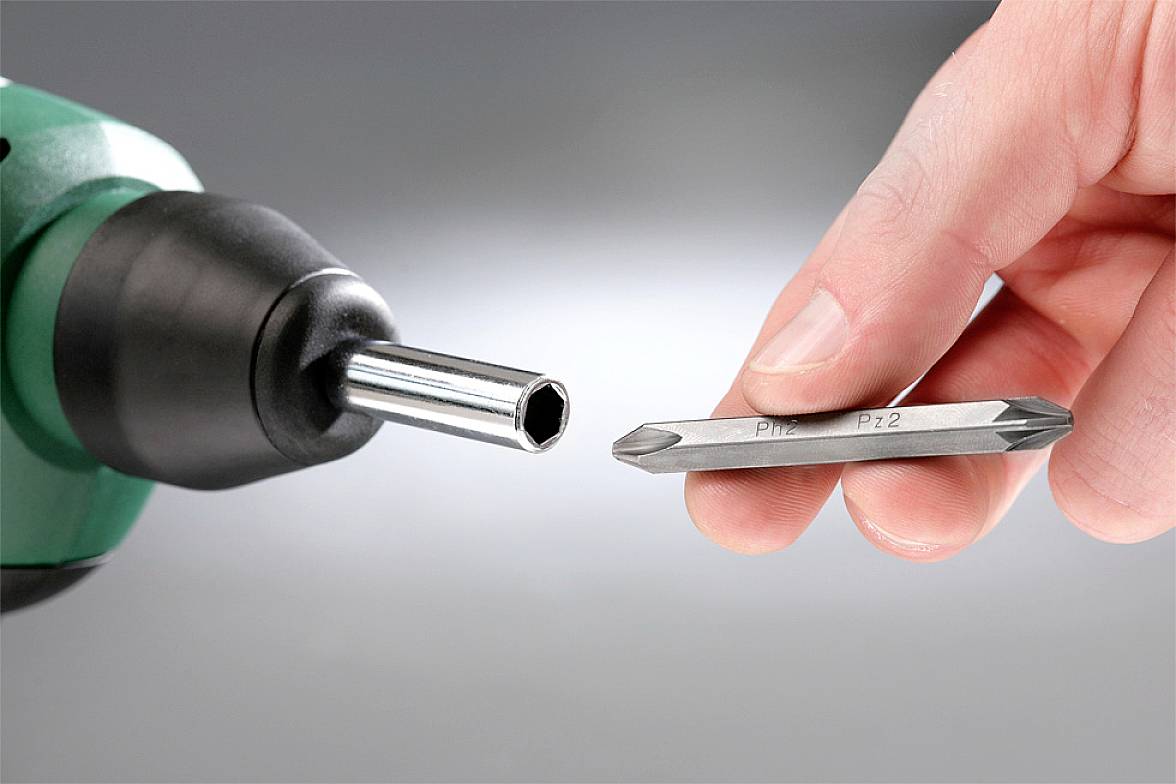A hand is holding a screwdriver bit in front of a cordless drill. The bit has two tips and is intended for assembly.