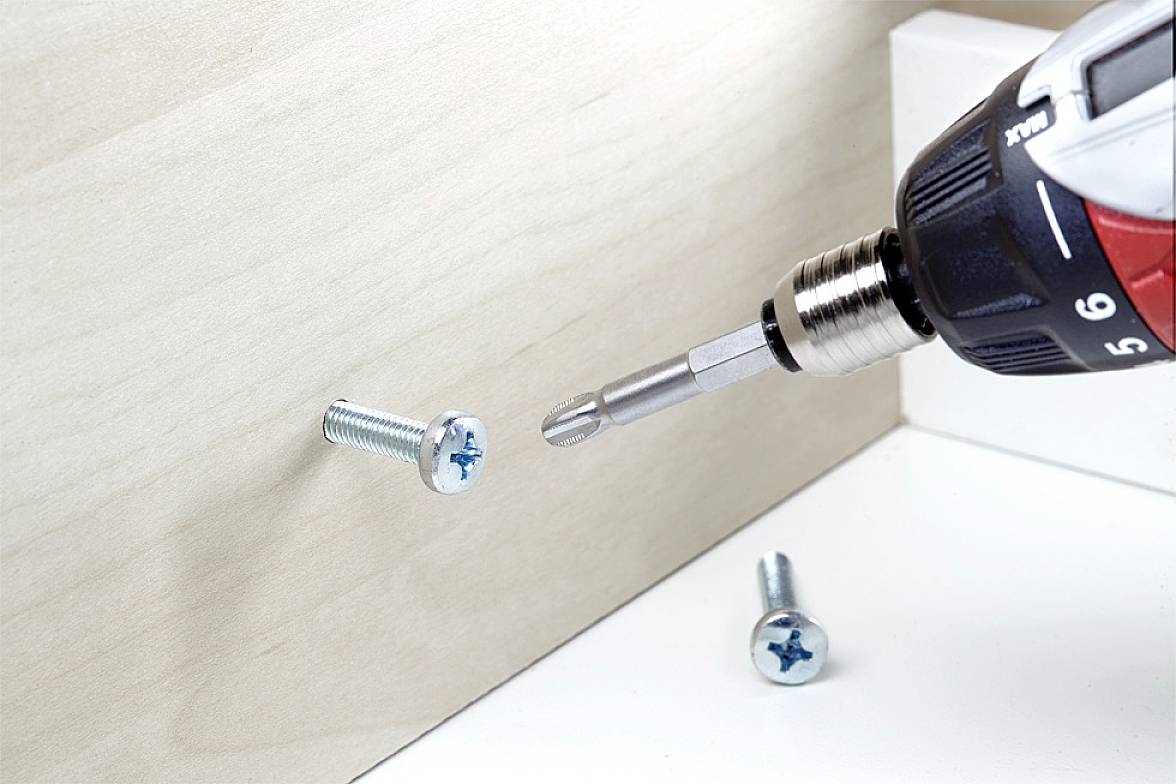 Cordless drill driving a screw into a wooden board. Another screw lies beside it.