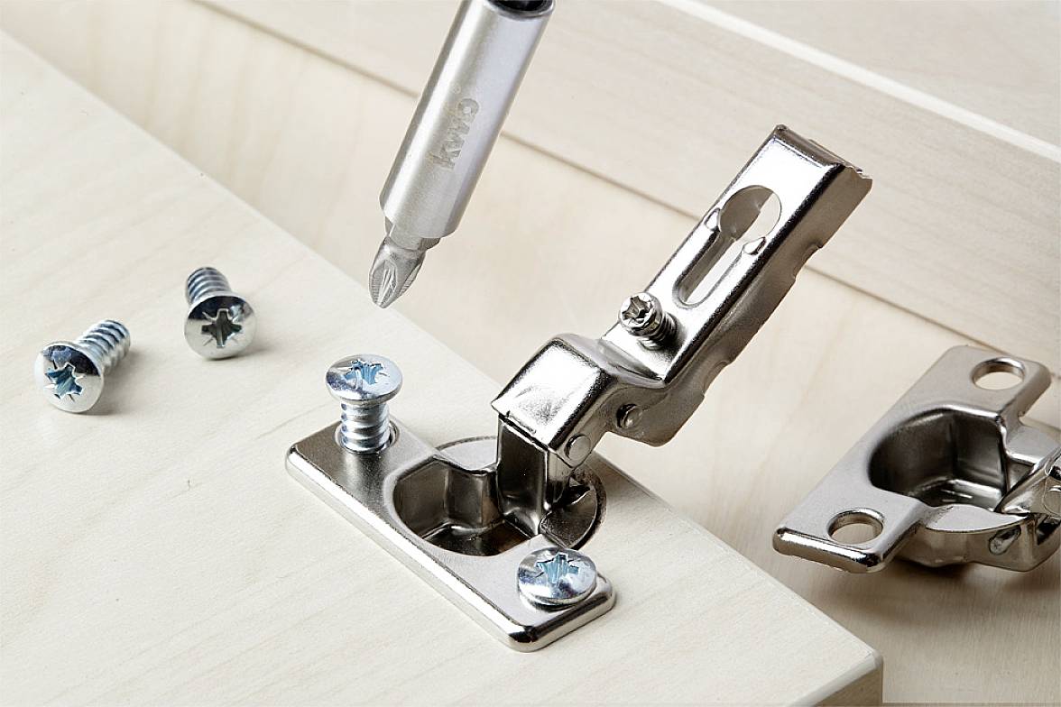 Metal hinge being attached to wooden furniture with a screwdriver. Two screws are laid out, with another hinge in the background.