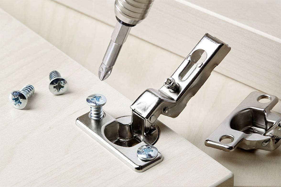 Close-up of a hinge on wood being secured with a screwdriver. Two additional screws are lying beside it.
