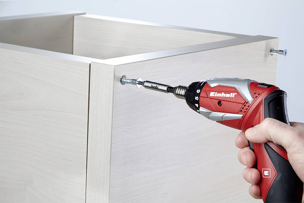 A person is using a red cordless drill to drive a screw into a wooden piece of furniture.