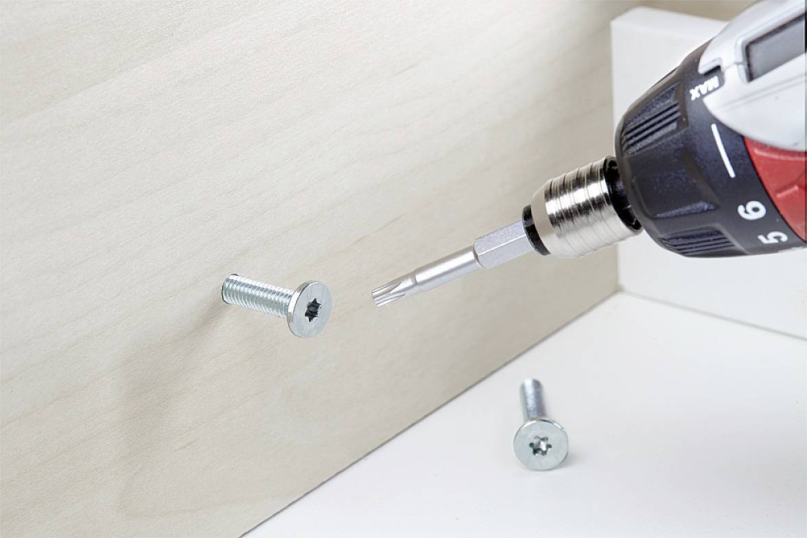 A drill with a screwdriver attachment is driving a screw into a wooden board. A second screw lies beside it.