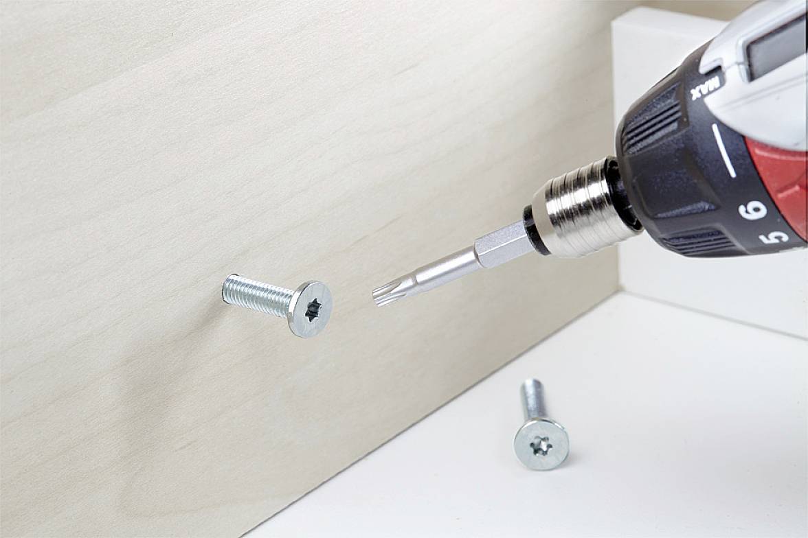 Power drill driving a screw into a piece of wood. Another screw lies nearby. The scene depicts furniture assembly or repair.