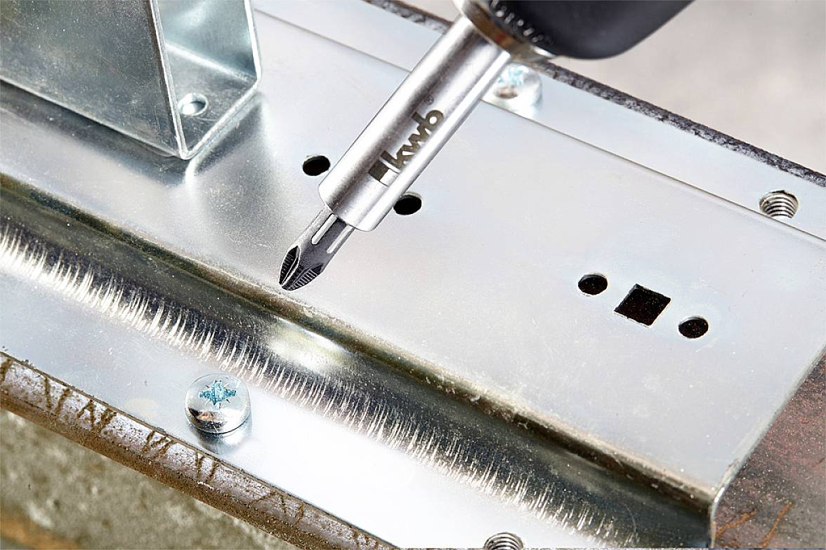 A screwdriver is driving a screw into a metal plate. The plate has several holes and is mounted on a support.