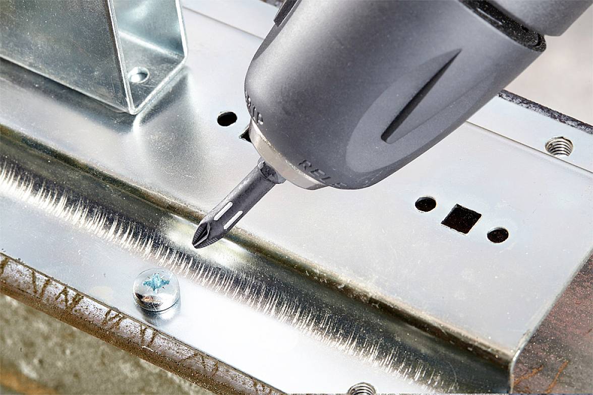 'Electric screwdriver drives in screw.' An electric power tool is used to precisely screw a screw into a piece of metal.