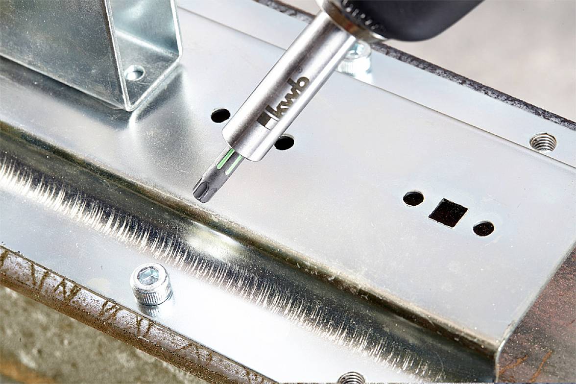 A power drill is tightening a metal plate with screws. The drill bit is pointing directly downwards, aimed at a gleaming surface.