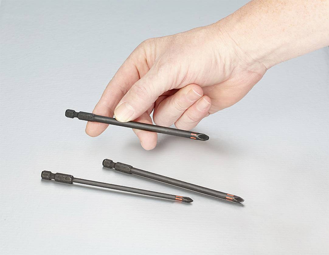 A hand holds a black screwdriver bit over a grey background. Two additional bits are lying on the surface.