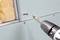 A drill drives a screw that fastens a green plasterboard sheet to a metal frame, with insulation material beside it.